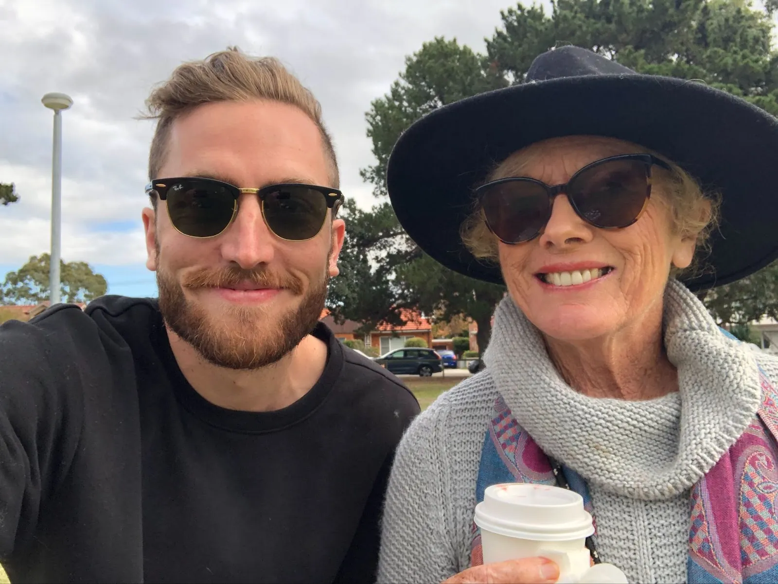 Man with sunglasses and woman in hat holding coffee smiling outdoors - Andy Crebar