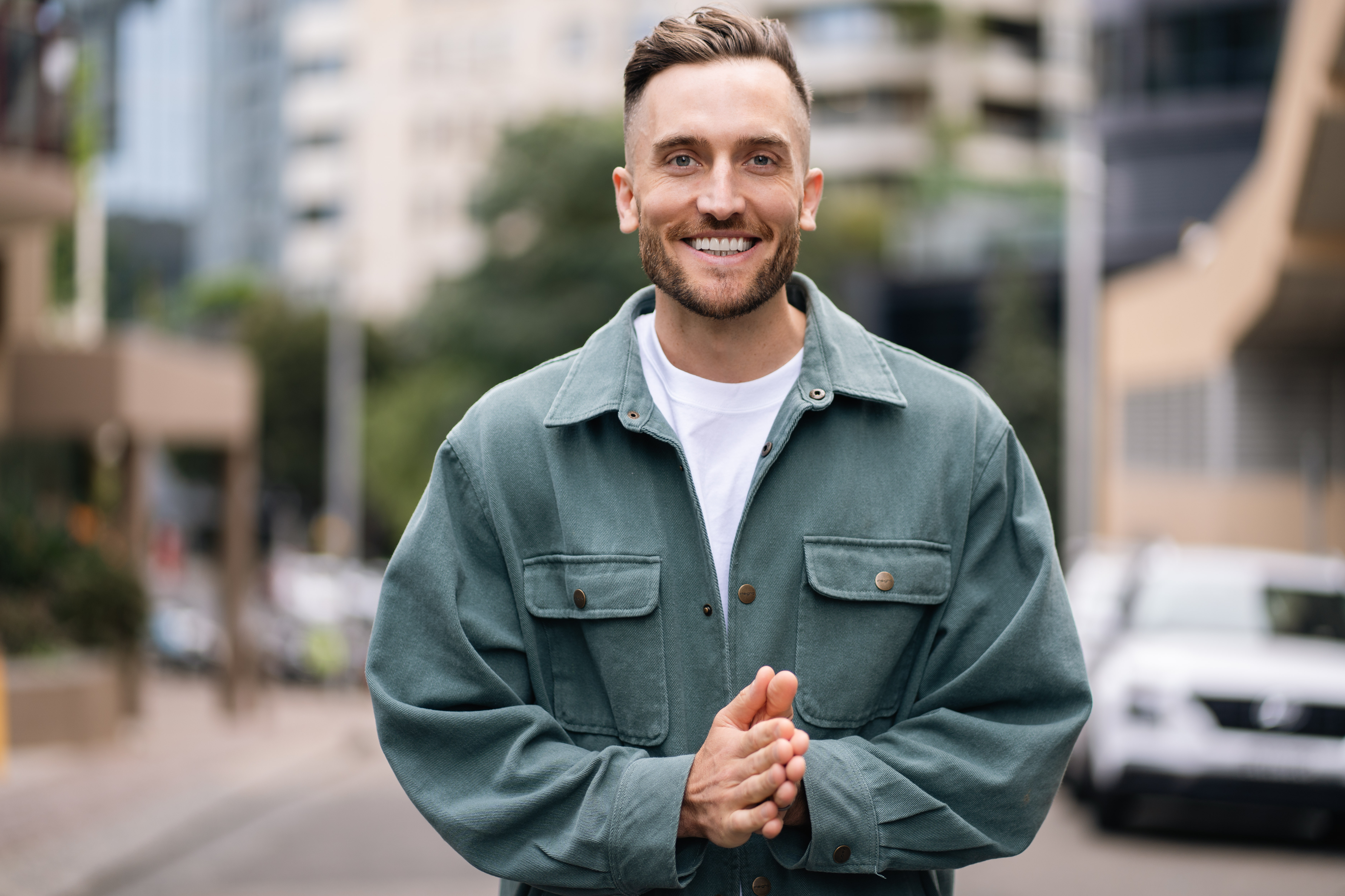 Andy Crebar standing on an urban street wearing a green jacket with hands clasped, smiling